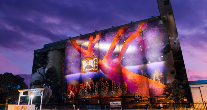 a colourful illustration of a couple and their car standing outdoors projected onto Karoonda Silo at dusk