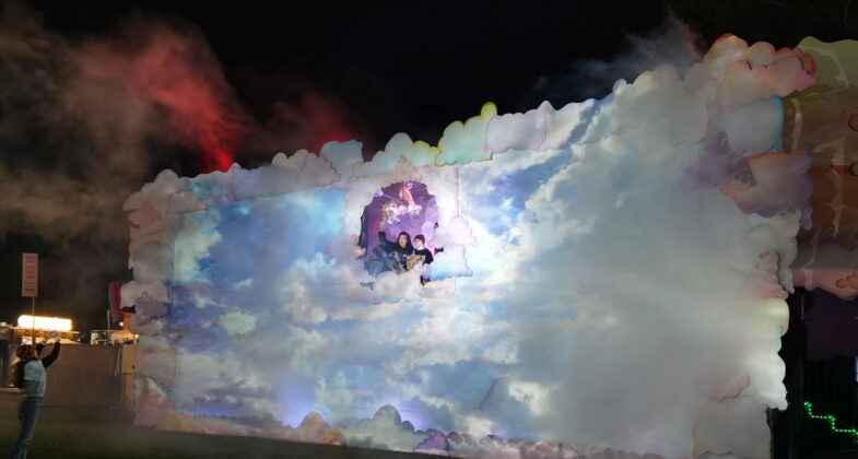 People enjoy a photo opportunity framed by a large illuminated scene with a cloudy glowing sky while a figure standing several feet below takes their picture.