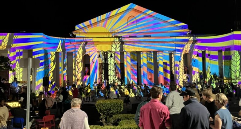 Moree Town Hall bathed in a vivid rainbow sunburst projection as crowd mingles.